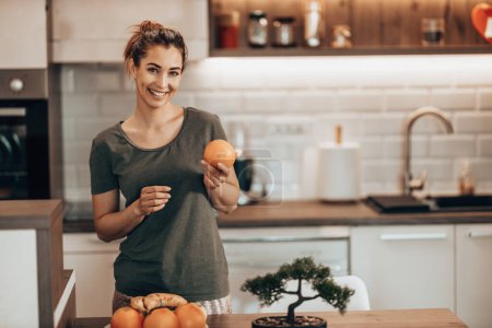 Téléchargez les photos : Tourné d'une jeune femme heureuse préparant un petit déjeuner de fruits dans sa cuisine à la maison. Regardant la caméra. - en image libre de droit