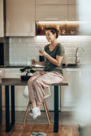 Photo for Young smiling woman using her smart phone while drinking coffee in the morning at home - Royalty Free Image