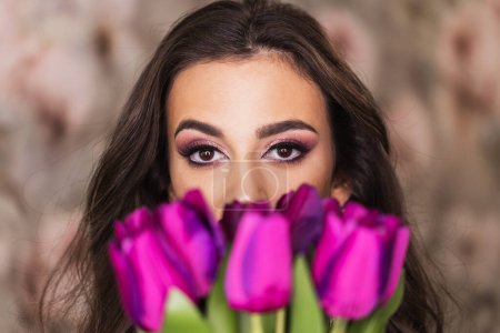 Foto de Un retrato de una joven atractiva con hermosos ojos, que sostiene un ramo de tulipanes morados y representa un bonito maquillaje en su cara. - Imagen libre de derechos