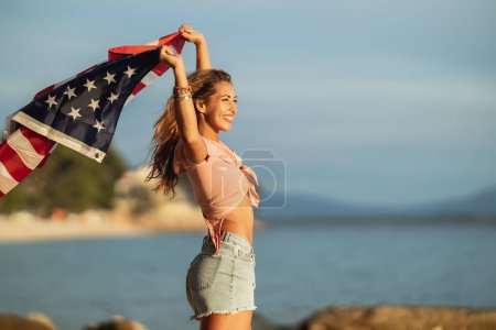 Foto de Mujer joven sonriente con bandera nacional americana disfrutando de un día relajante en la playa. - Imagen libre de derechos