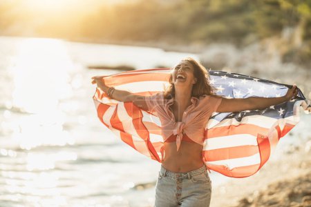 Foto de Happy young woman with American national flag having fun and spending the day on the beach. - Imagen libre de derechos