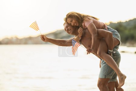 Foto de Una pareja alegre y cariñosa con bandera nacional de Estados Unidos divirtiéndose mientras pasa el día en la playa. - Imagen libre de derechos