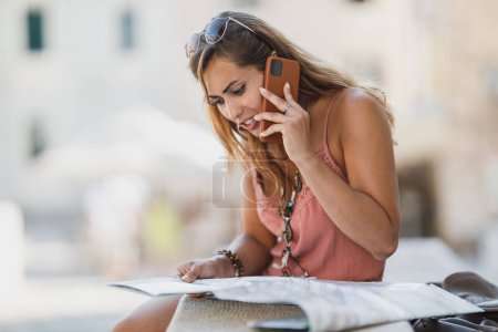 Foto de Mujer hablando en un teléfono inteligente mientras está sentada en las escaleras de la calle con mapa en las manos y explorando una ciudad mediterránea. - Imagen libre de derechos