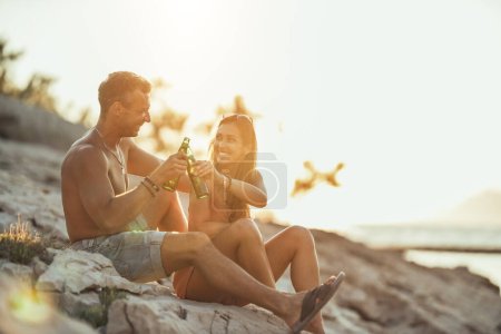 Téléchargez les photos : Young couple having fun and drinking beer on the beach by the sea. Enjoying in beautiful sunset and their love. - en image libre de droit
