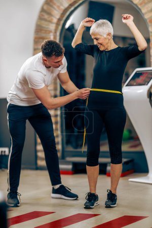 Téléchargez les photos : Instructeur de fitness mesurant la taille de la femme âgée avant de s'entraîner dans la salle de gym. - en image libre de droit