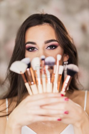 Photo for A portrait of a cute young woman with beautiful eyes, who is holding makeup brushes and representing a nice makeup on her face. - Royalty Free Image