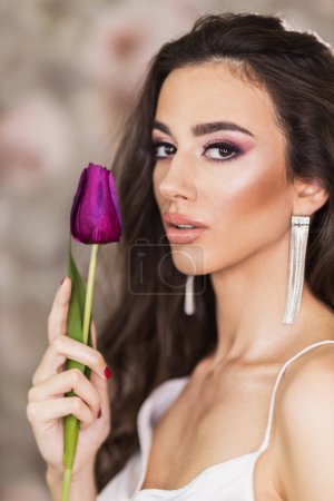 Foto de Un retrato de una linda joven que representa un bonito maquillaje en su cara y sostiene un tulipán púrpura. Mirando la cámara. - Imagen libre de derechos