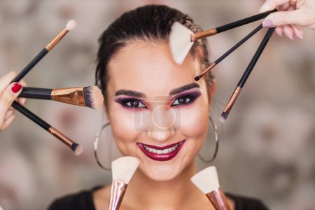 Photo for A portrait of a cute young woman surrounded by makeup brushes who is presenting a nice makeup on her face. - Royalty Free Image