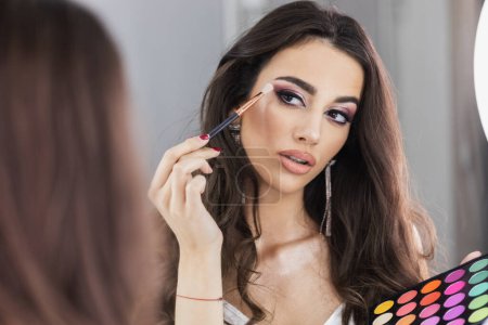 Photo for Beautiful young woman is applying eyeshadow in front of mirror. Focus on the reflection in the mirror. - Royalty Free Image