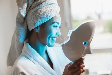 Foto de Hermosa mujer joven recibiendo un tratamiento de máscara de terapia de luz led para su cara en el salón de belleza. - Imagen libre de derechos