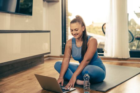 Téléchargez les photos : Jeune femme utilisant un ordinateur portable tout en faisant de la formation de conditionnement physique à la maison le matin. - en image libre de droit