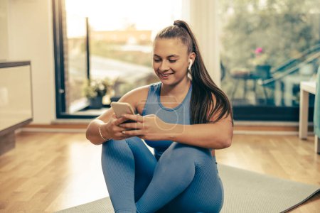 Photo for Young smiling woman listening music from her smartphone and preparing for training at home in the morning. - Royalty Free Image