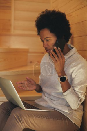 Foto de Mujer negra hablando en un teléfono inteligente mientras usa un ordenador portátil durante las vacaciones en una acogedora casa de madera en un bosque. - Imagen libre de derechos