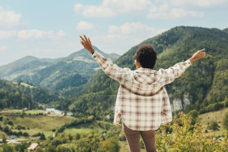 Foto de Vista trasera de una mujer negra con los brazos extendidos de pie en la naturaleza y disfrutando al aire libre. - Imagen libre de derechos