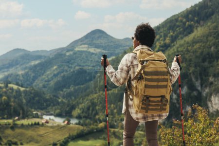 Foto de Vista trasera de una mujer negra con mochila y bastones de trekking de pie en la naturaleza y disfrutando al aire libre mientras camina por la montaña. - Imagen libre de derechos