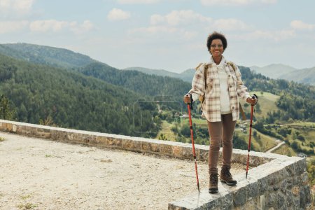 Foto de Mujer negra madura con mochila y bastones de trekking de pie en la naturaleza y disfrutando al aire libre mientras camina por la montaña. - Imagen libre de derechos