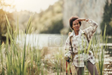 Photo for Mature black woman with backpack standing by the lake  relaxing from his hiking activity in nature. - Royalty Free Image
