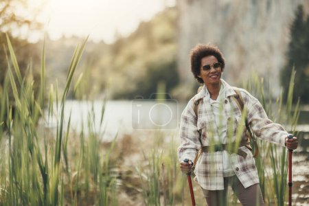 Photo for Relaxed mature black woman with backpack and trekking poles  standing by the river and relaxing while out hiking on mountain. - Royalty Free Image