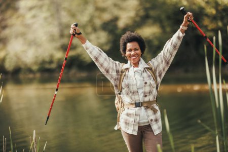 Téléchargez les photos : Femme noire mature avec les bras tendus tenant des bâtons de trekking et profitant au bord du lac pendant la randonnée en montagne. - en image libre de droit