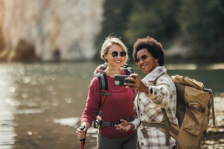 Photo for Two mixed-race female friends taking selfies by a lake during a hike along the mountain. - Royalty Free Image