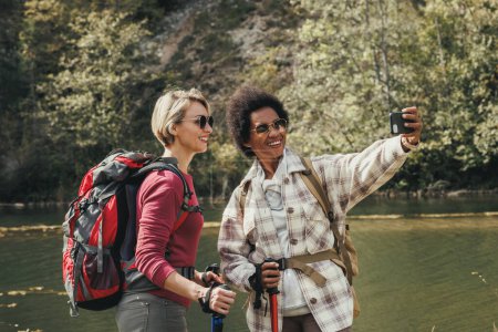 Téléchargez les photos : Deux amies métisses prenant des selfies au bord d'une rivière lors d'une randonnée le long de la montagne. - en image libre de droit