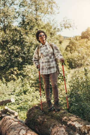 Téléchargez les photos : Femme noire mature avec sac à dos et bâtons de trekking profitant de la vue sur la forêt pendant la randonnée dans la nature. - en image libre de droit