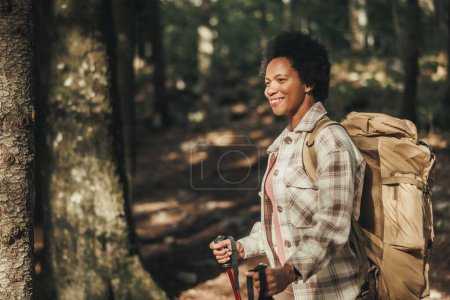 Foto de Sonriente mujer negra madura parada sola afuera y llevando una mochila mientras caminaba en las montañas. - Imagen libre de derechos