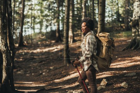 Téléchargez les photos : Femme noire mature avec sac à dos profitant de la vue sur la forêt pendant la randonnée dans la nature. - en image libre de droit