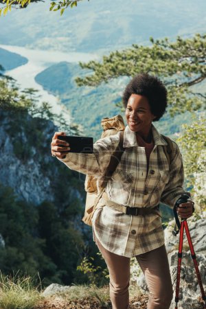Téléchargez les photos : Femme noire mature faire un selfie tout en se tenant sur le sommet d'une colline et profiter de la vue lors de sa randonnée dans les montagnes. - en image libre de droit