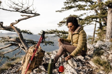 Téléchargez les photos : Femme noire mature assise sur le sommet d'une colline et faisant une pause thé pendant sa randonnée dans les montagnes. - en image libre de droit
