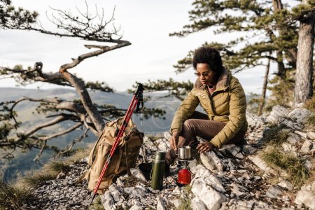 Téléchargez les photos : Femme noire mature assise sur le sommet d'une colline et faisant une pause thé pendant sa randonnée dans les montagnes. - en image libre de droit