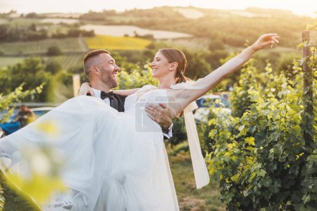 Foto de Alegre novio llevando y girando a su novia mientras disfruta en el viñedo el día de su boda. - Imagen libre de derechos