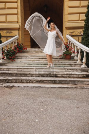 Foto de Hermosa novia divirtiéndose y bailando por las escaleras al aire libre mientras su largo velo nupcial volando en el aire. - Imagen libre de derechos