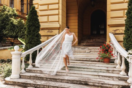 Photo for Beautiful bride walking down the stairs outdoor while her bridal long veil flying on wind in air. - Royalty Free Image