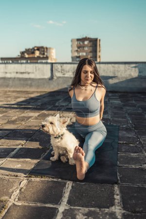 Photo for Relaxed woman practicing yoga stretching exercise on a rooftop supporting by her pet dog. - Royalty Free Image