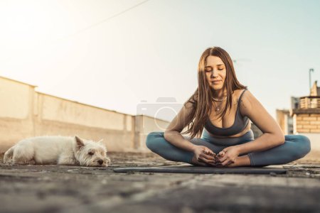Téléchargez les photos : Relaxed woman practicing yoga on a rooftop terrace at sunset supporting by her pet dog. - en image libre de droit