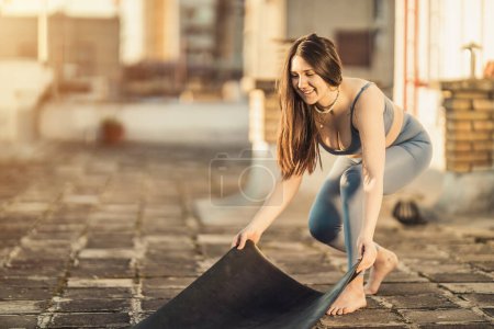 Téléchargez les photos : Femme détendue prépare tapis d'exercice pour pratiquer le yoga sur un toit-terrasse au coucher du soleil. - en image libre de droit