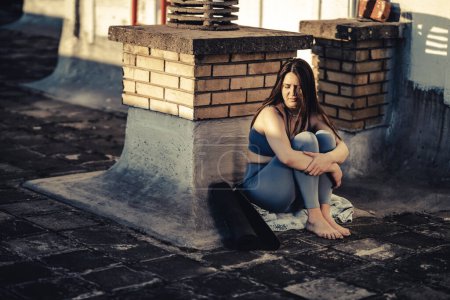 Téléchargez les photos : Jeune femme seule dans la dépression s'asseoir avec embrasser les genoux sur une terrasse sur le toit perdu dans de mauvaises pensées pessimistes après la formation de yoga. - en image libre de droit