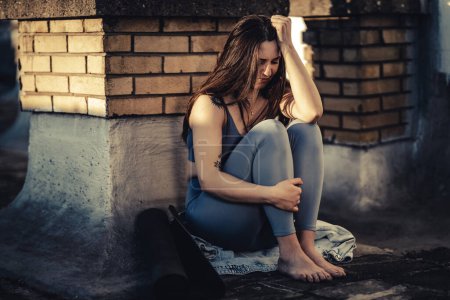 Téléchargez les photos : Jeune femme en dépression assis sur un toit-terrasse perdu dans de mauvaises pensées pessimistes et se préparer à l'entraînement en plein air. - en image libre de droit