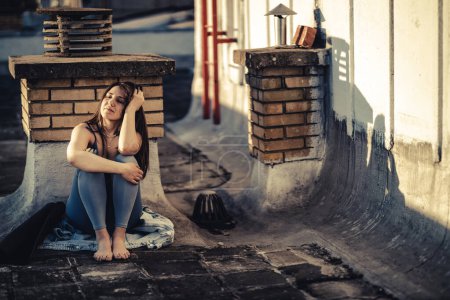 Téléchargez les photos : Jeune femme seule dans la dépression s'asseoir avec embrasser les genoux perdus dans de mauvaises pensées pessimistes et se préparer à l'entraînement sur un toit-terrasse. - en image libre de droit