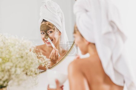 Téléchargez les photos : Belle jeune femme devant le miroir appliquant un masque facial dans la salle de bain à la maison. - en image libre de droit