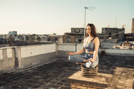 Téléchargez les photos : Femme détendue pratiquant le yoga méditant sur un toit-terrasse. Bâtiments municipaux en arrière-plan. - en image libre de droit
