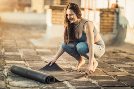 Foto de La mujer relajada está enrollando la esterilla de ejercicio y preparándose para practicar yoga en una terraza en la azotea al atardecer.. - Imagen libre de derechos