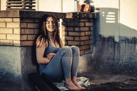 Téléchargez les photos : Jeune femme pensant à quelque chose tout en s'asseyant sur un toit-terrasse après l'entraînement de yoga au coucher du soleil. - en image libre de droit
