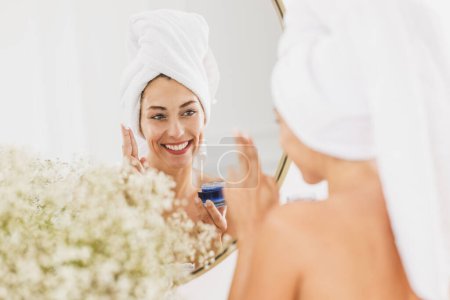 Photo for Beautiful young woman in front of mirror applying moisturizer on her face in the bathroom at home. - Royalty Free Image