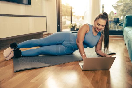 Photo for Young woman using laptop while doing fitness exercises at home in the morning. - Royalty Free Image