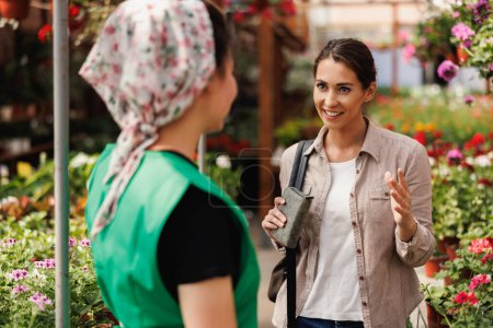 Photo for Young female plant nursery volunteer helps a young woman with flowers choosing and purchase. - Royalty Free Image
