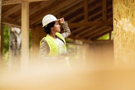 Téléchargez les photos : Prise de vue d'une architecte afro-américaine vérifiant le chantier d'une nouvelle maison en bois. Elle porte des vêtements de travail protecteurs et un casque blanc. - en image libre de droit