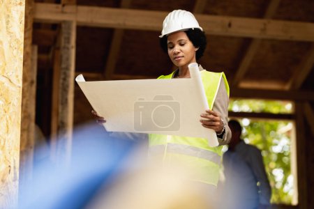 Photo for Shot of an African American female architect checking blueprints at the construction site of a new wooden house. She is wearing protective workwear and white helmet. - Royalty Free Image