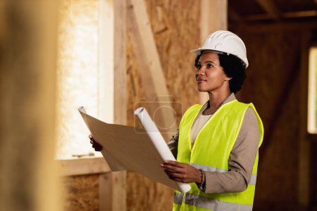 Foto de Fotografía de una arquitecta afroamericana revisando planos en el sitio de construcción de una nueva casa de madera. Ella está usando ropa de trabajo protectora y casco blanco. - Imagen libre de derechos